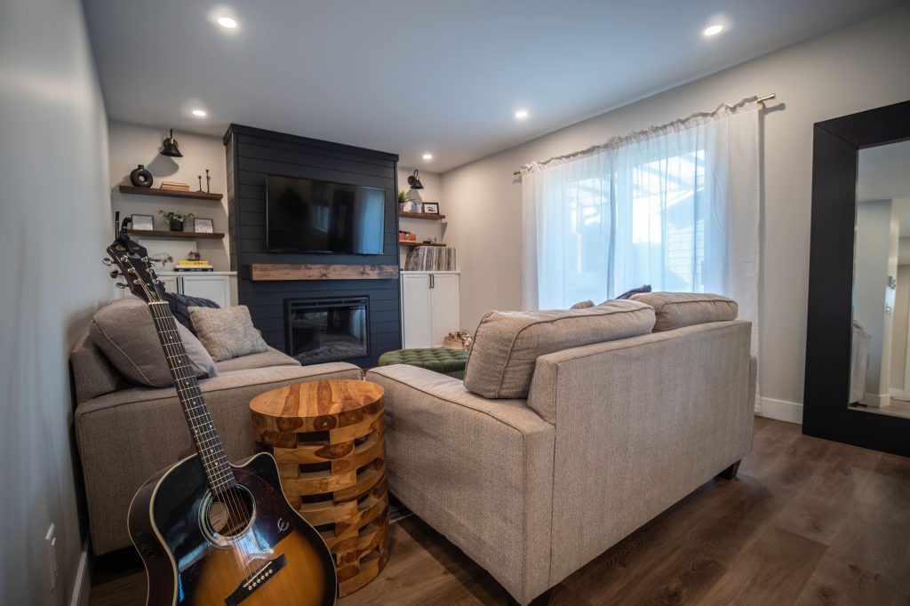 Cozy living room featuring a modern design with a gray sofa, wooden accent table, acoustic guitar, wall-mounted TV, and natural light from sheer curtains, showcasing DMC Contracting's custom home craftsmanship.