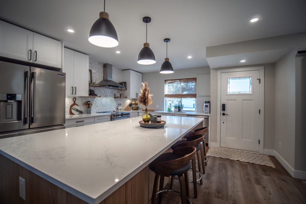 Modern kitchen renovation featuring a spacious island with bar stools, stainless steel appliances, white cabinetry, and stylish pendant lighting, showcasing quality craftsmanship by DMC Contracting in a bright, inviting space.