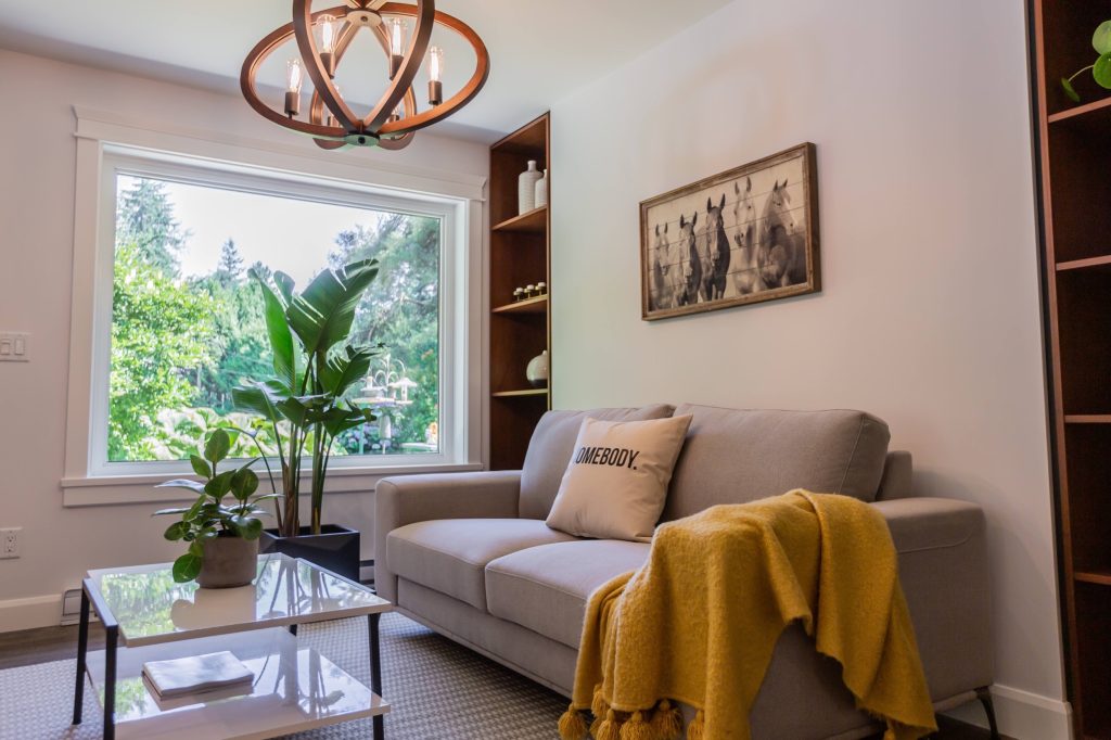 Bright living room featuring a modern gray sofa with a decorative pillow, yellow throw blanket, glass coffee table, and large window showcasing lush greenery, emphasizing custom design and comfort in luxury renovations.