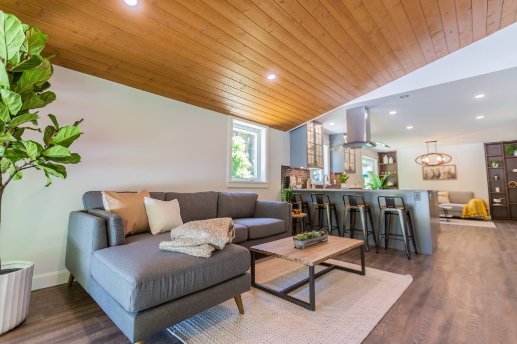 Modern living room with gray sectional sofa, wooden ceiling, and open kitchen featuring bar stools, showcasing a blend of comfort and contemporary design for home renovation inspiration in the Fraser Valley.