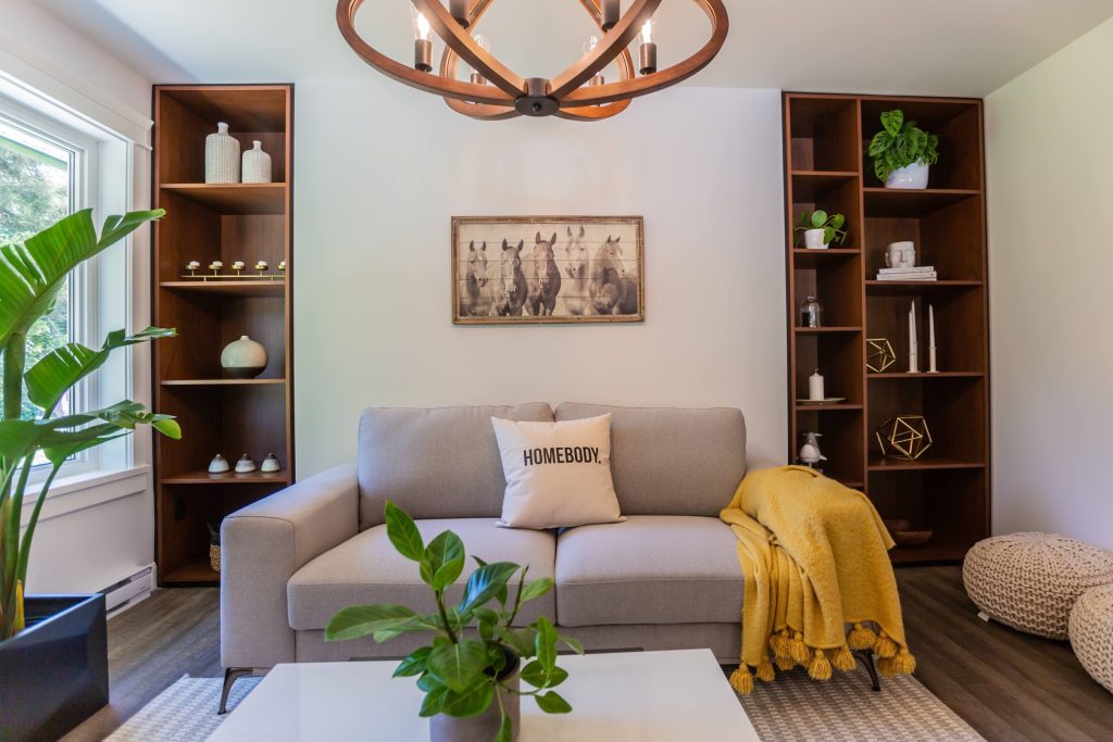 Cozy living room with a gray sofa adorned with a "HOMEBODY" pillow, yellow throw blanket, decorative plants, and stylish shelving, reflecting adaptable and inviting design for future-proof homes.