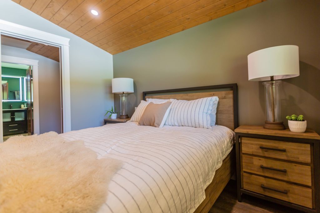 Cozy bedroom featuring a wooden bed with striped bedding, bedside tables with lamps, and a view into an adjoining bathroom, reflecting multi-generational living design for comfort and privacy.