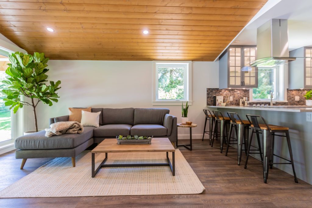 Modern living room with gray sectional sofa, wooden ceiling, and kitchen bar area, showcasing custom home design by DMC Contracting in Abbotsford.