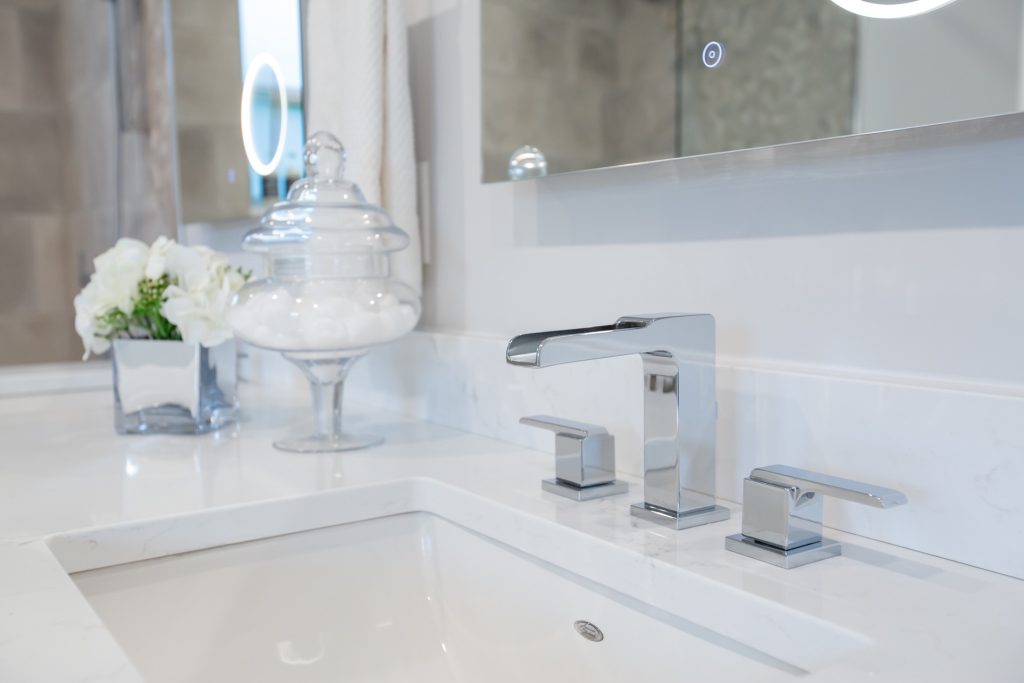 Luxury bathroom sink with modern chrome faucets, elegant white marble countertop, glass jar with cotton balls, and fresh white flowers in a silver vase, reflecting high-end finishes in a renovated space.