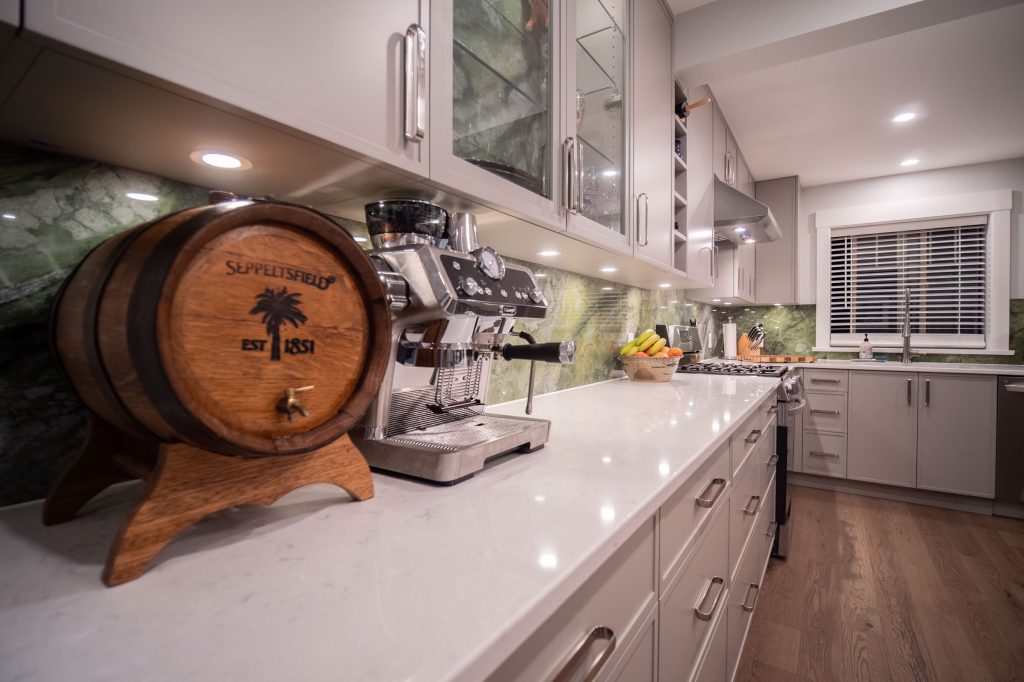 Modern kitchen featuring sleek cabinetry, a high-end espresso machine, and a decorative wooden barrel, complemented by a green stone backsplash and natural light from a window.