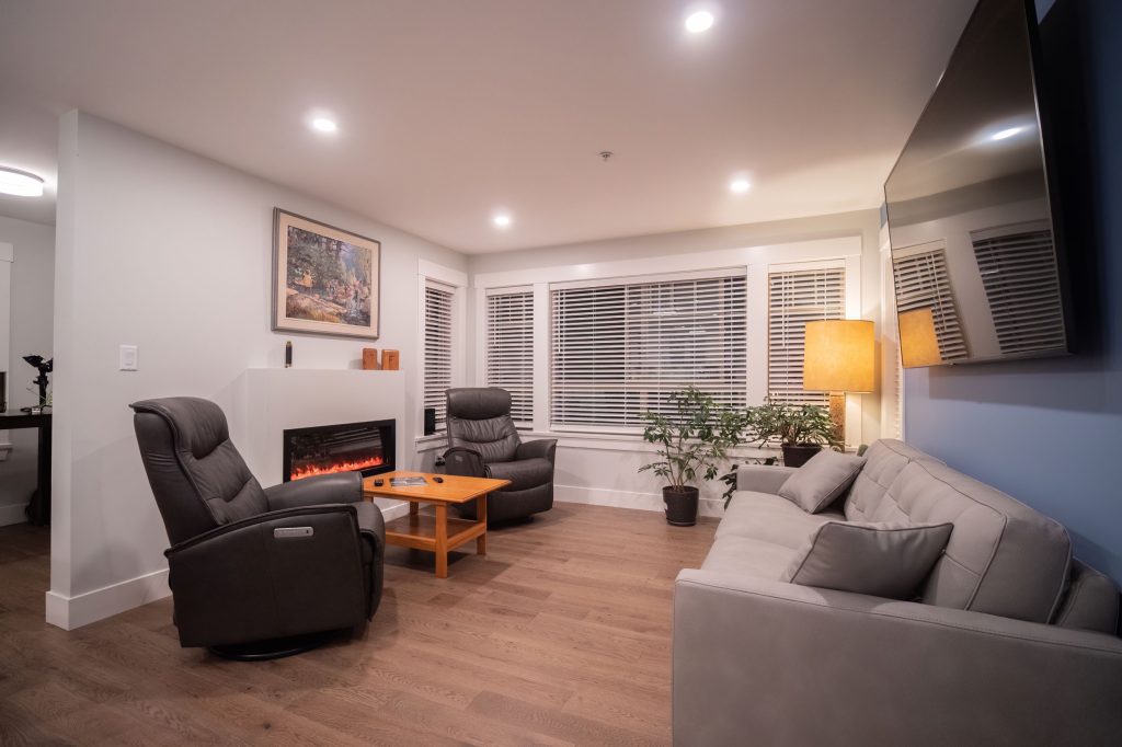 Modern living room with gray sofa, recliner chairs, wooden coffee table, and a cozy fireplace, featuring large windows with blinds, stylish lighting, and decorative plants, illustrating a renovated space for comfort and efficiency.