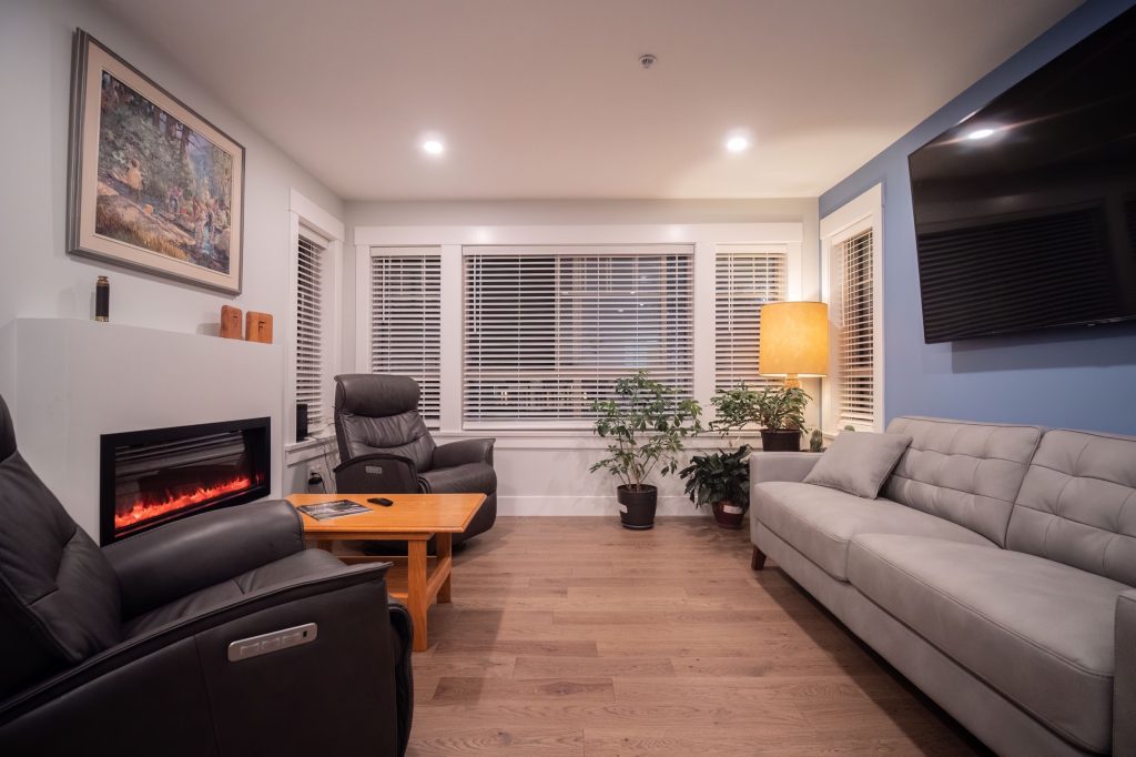 Modern living room with sleek gray sofa, black recliner chairs, electric fireplace, wooden coffee table, and indoor plants, showcasing a stylish and comfortable renovation space.