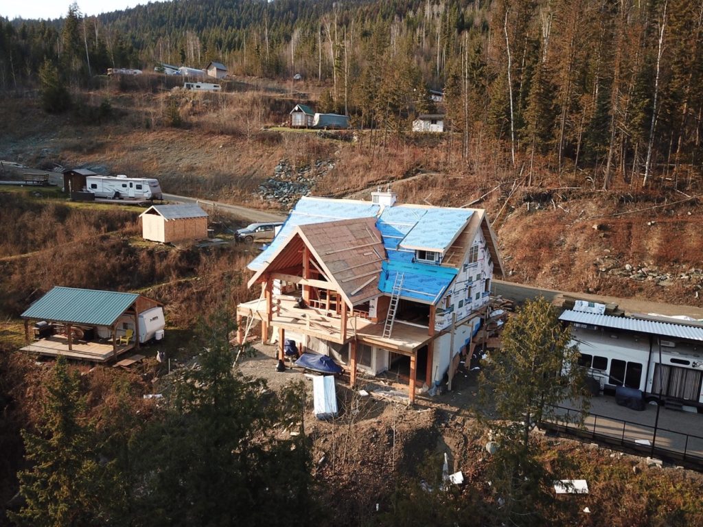 Custom home under construction in a scenic rural setting, featuring wooden framing, a blue tarp roof, and nearby structures, emphasizing DMC Contracting's expertise in building tailored homes in the Fraser Valley.