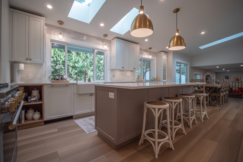 Modern kitchen design featuring white cabinetry, large windows with greenery view, and stylish pendant lighting over a spacious island with seating, emphasizing custom home aesthetics and functional living spaces in Fraser Valley.