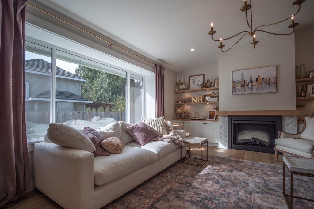 Modern living room featuring a light-colored sofa with decorative pillows, large windows for natural light, stylish chandelier, and elegant shelving with decor, emphasizing a renovated and inviting space ideal for family gatherings in Abbotsford.