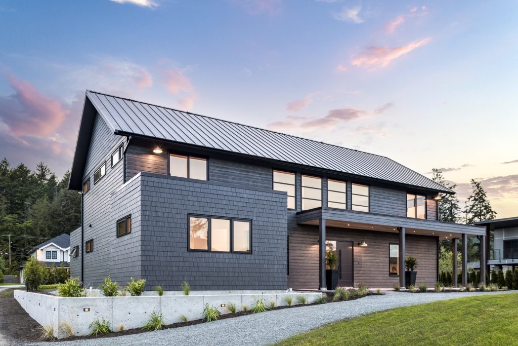 Modern custom home design with dark wood siding, large windows, and a metal roof, set in a landscaped yard in the Fraser Valley.